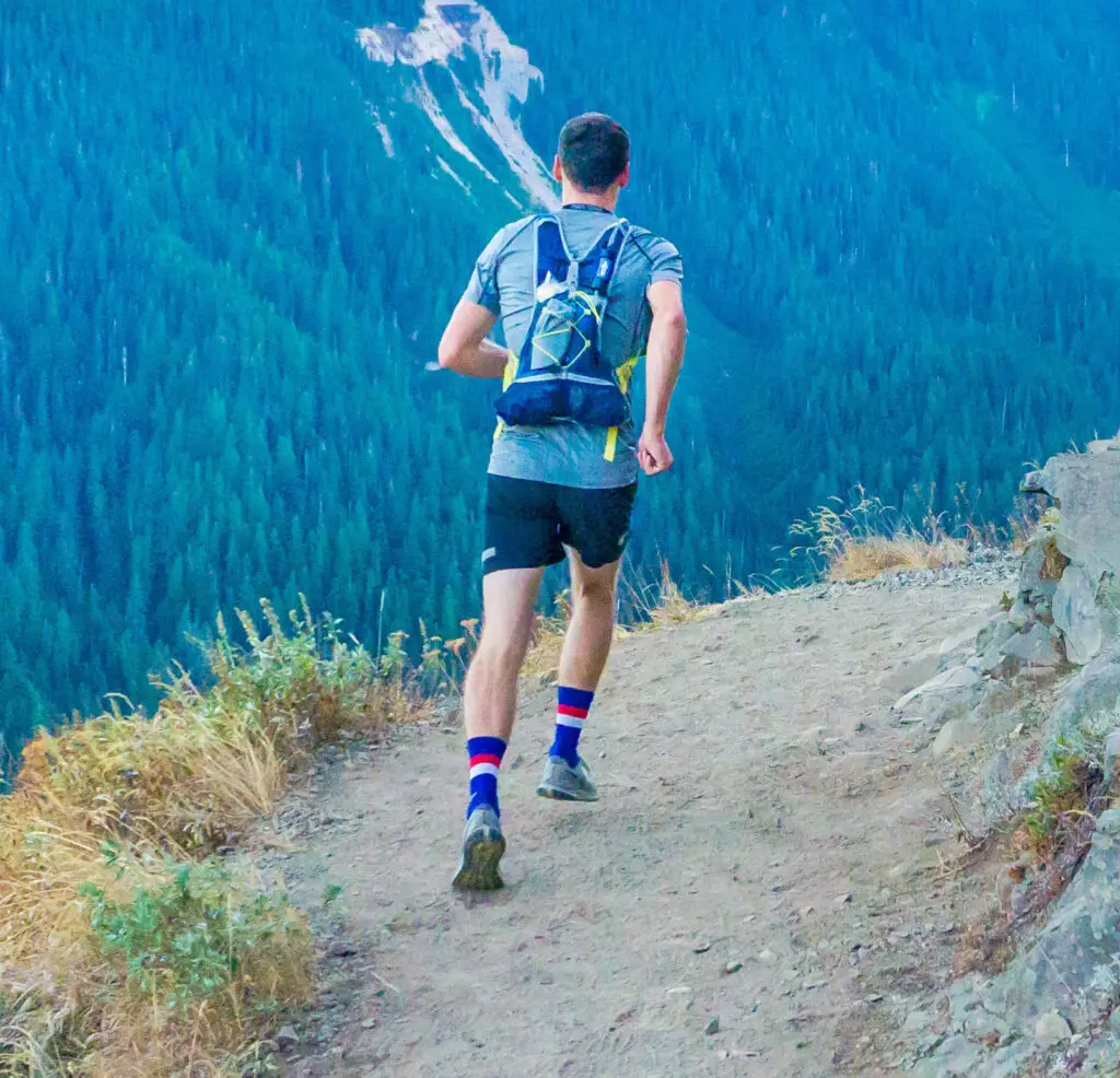 Person läuft auf einem schmalen Pfad am Berg entlang, umgeben von dichter Waldlandschaft; trägt Sportbekleidung und einen Rucksack.