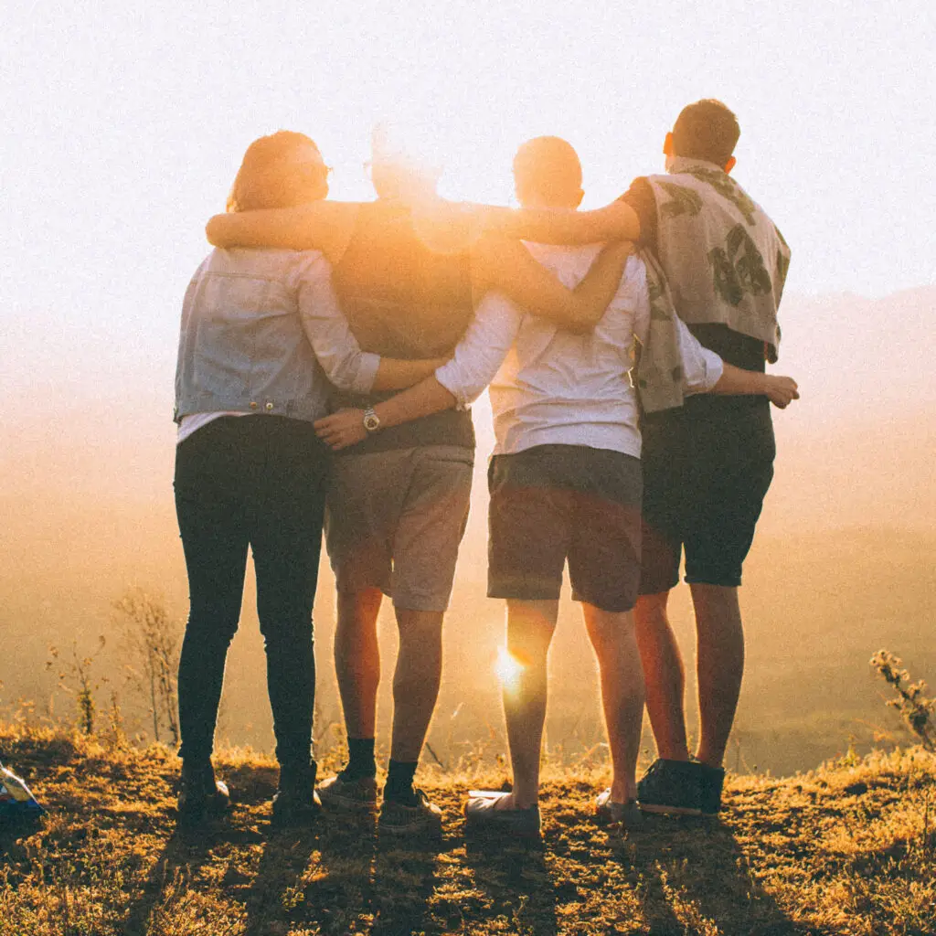 Vier Personen umarmen sich und stehen auf einer Wiese bei Sonnenuntergang, die Sonne scheint zwischen ihnen hindurch. Sie blicken in die Ferne und tragen legere Kleidung.
