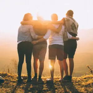 Vier Personen umarmen sich und stehen auf einer Wiese bei Sonnenuntergang, die Sonne scheint zwischen ihnen hindurch. Sie blicken in die Ferne und tragen legere Kleidung.