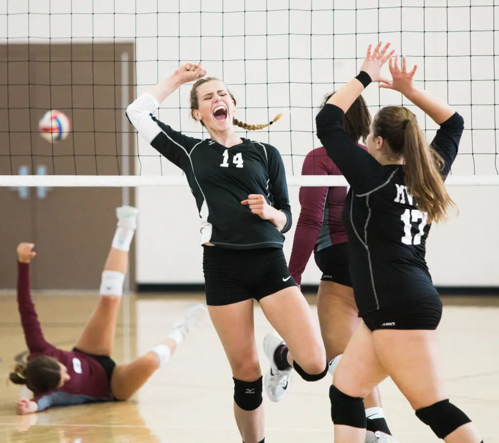 Volleyballspiel in einer Sporthalle, zwei Spielerinnen in schwarzen Trikots mit den Nummern 14 und 17 jubeln vor dem Netz, während im Hintergrund eine Spielerin in einem weinroten Trikot auf dem Boden liegt. Der Ball ist auf dem Weg ins Aus. Dynamische Spielsituation.
