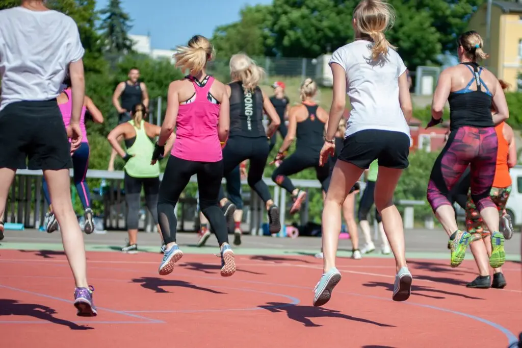 Gruppe von Menschen, hauptsächlich Frauen, die auf einem Sportplatz an einer Fitnessübung im Freien teilnehmen. Sie tragen Sportkleidung und springen synchron in die Luft. Im Hintergrund sind Bäume und Gebäude zu sehen.