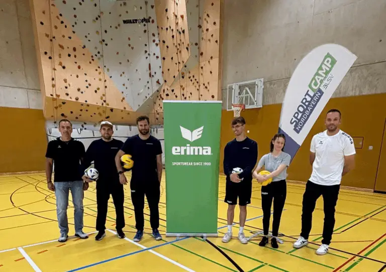 Sieben Personen stehen in einer Sporthalle auf einem gelb markierten Spielfeld. Sie halten bunte Bälle und stehen jeweils links und rechts neben zwei aufgestellten Werbebannern, einem für "Erima" und einem für "Sport-Camp Nordbayern". Im Hintergrund ist eine Kletterwand und ein Basketballkorb zu sehen.