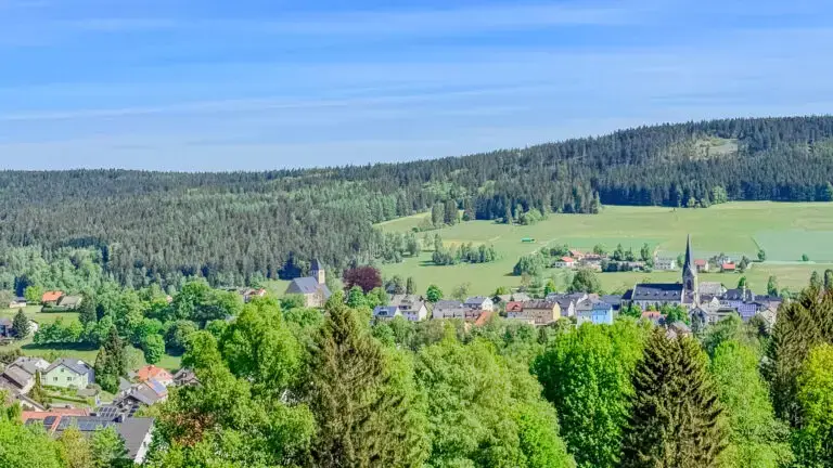Kleines Dorf in einer grünen Landschaft, umgeben von Wäldern. Im Vordergrund sind Häuser mit roten Dächern zu sehen, in der Mitte steht eine Kirche mit einem spitzen Turm. Der Hintergrund zeigt eine ausgedehnte Waldfläche unter einem blauen Himmel.