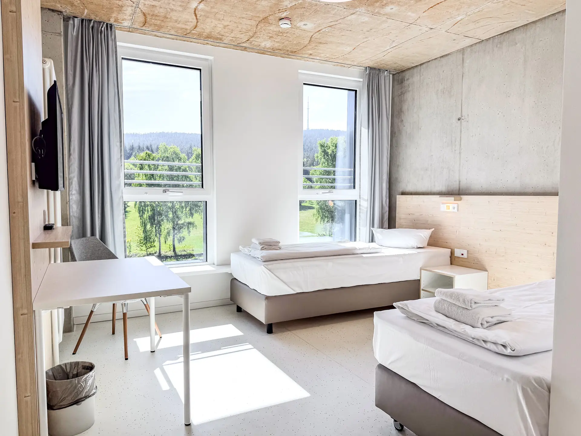 Heller Raum mit zwei Einzelbetten, hellem Holz und Betonwand. Große Fenster mit grauen Vorhängen bieten Blick auf eine grüne Landschaft. Ein Tisch und ein Stuhl stehen links neben einem Mülleimer. Ein Fernseher hängt an der Wand.
