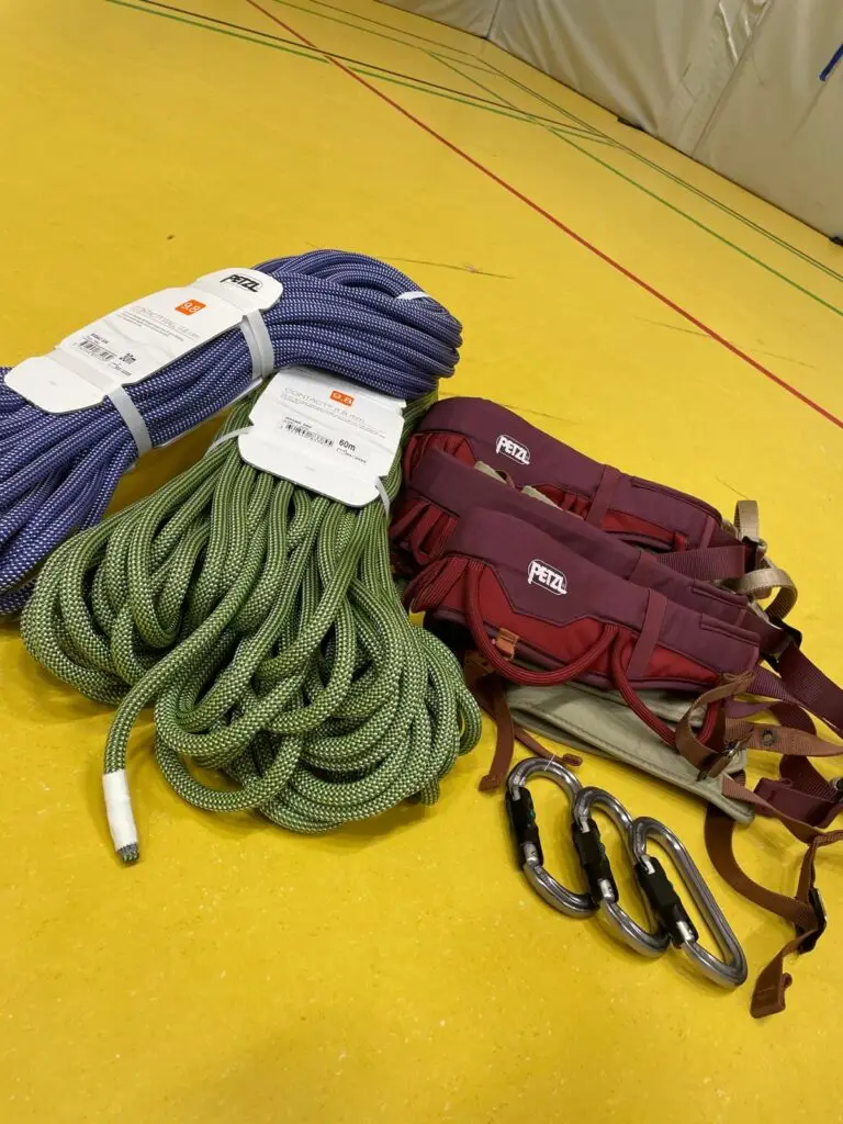 Kletterausrüstung auf gelbem Boden, einschließlich zwei Seile in Blau und Grün, zwei rote Klettergurte und drei silberne Karabiner.