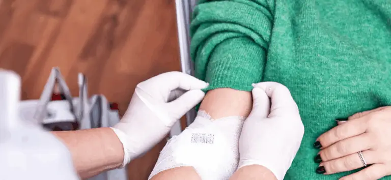 Eine Person im grünen Pullover erhält eine Blutspende oder Blutinjektion. Ein medizinisches Personal trägt weiße Handschuhe und legt den Arm der Person mit einem Verband an. Im Hintergrund sind Blutbeutel sichtbar.