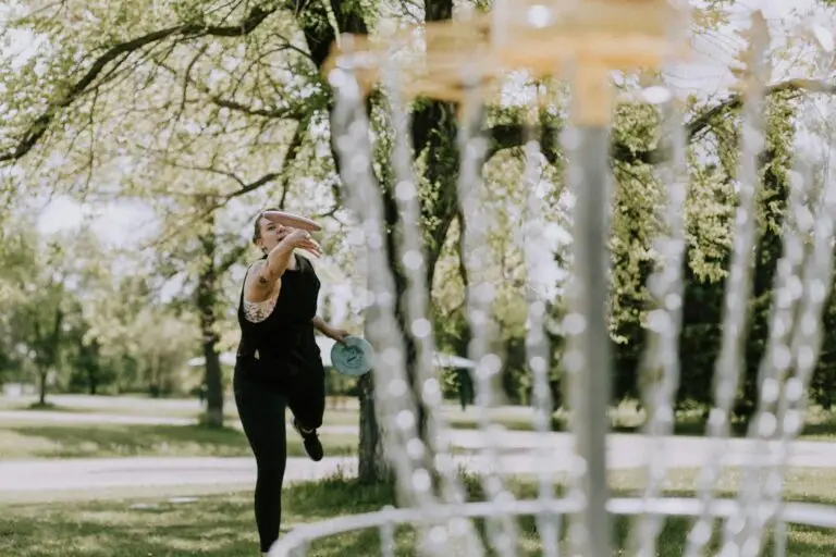 Person wirft eine Frisbee-Scheibe im Freien in Richtung eines Discgolf-Korbs. Bäume im Hintergrund, sonniges Wetter.