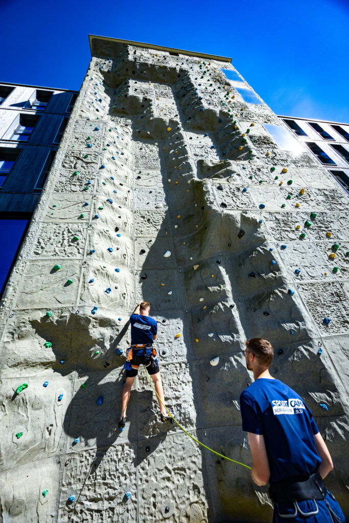 Ein Übungsleiter vom BLSV Sportcamp Nordbayern klettert an der Outdoor Kletterwand. Sein Kollege sichert ihn. Beide sind in den blauen Übungsleiter T-Shirts des Sportcamps.